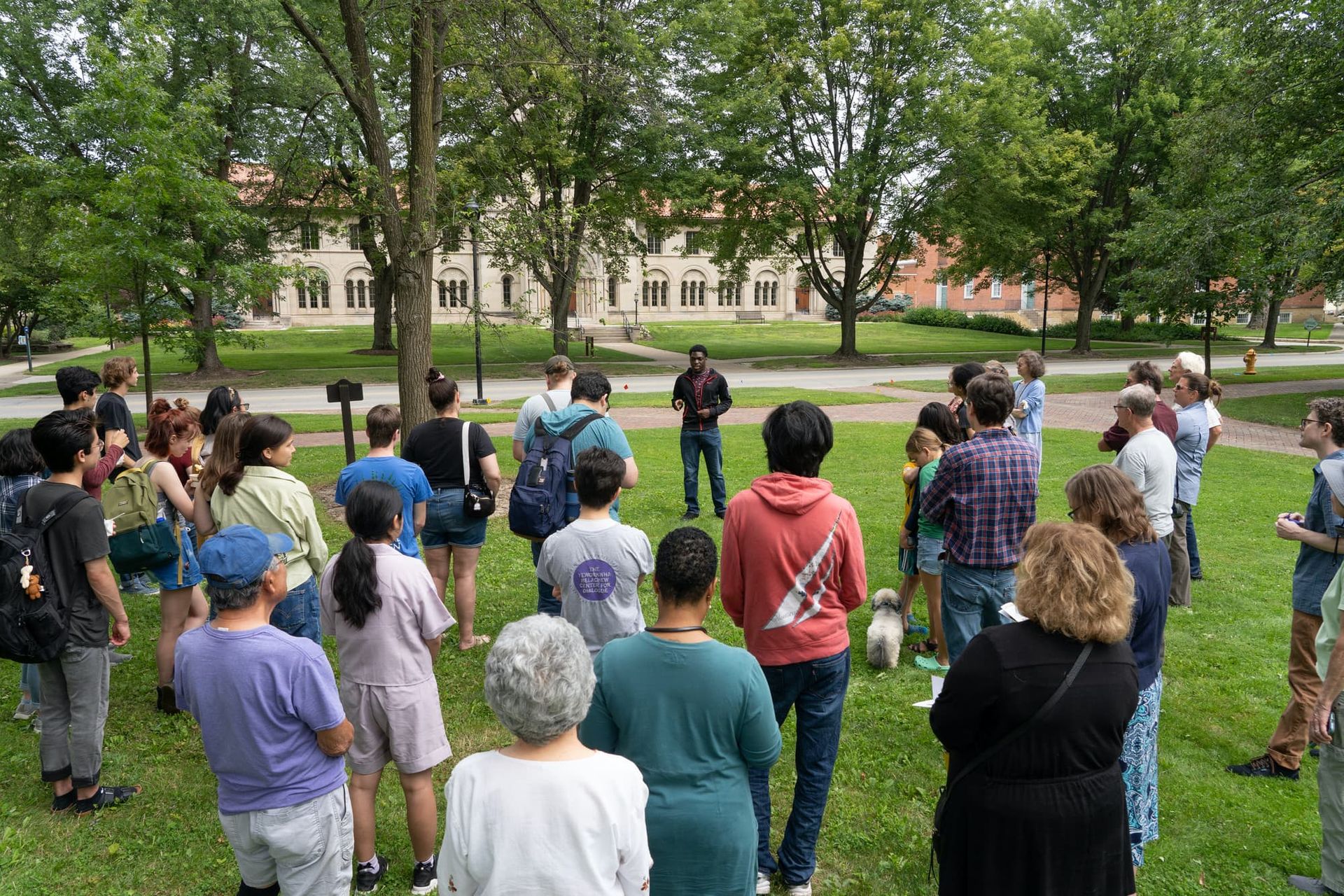 A group of people stand in a small parkland listen to a talk by a young speaker.