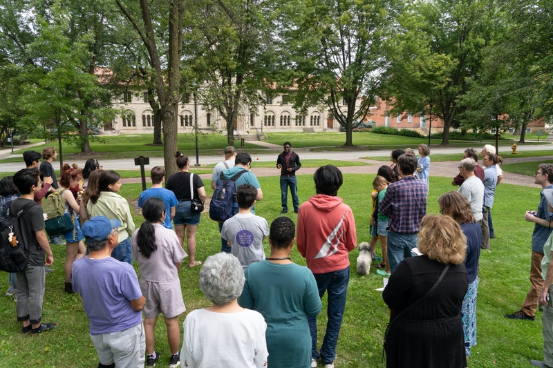 A group of people stand in a small parkland listen to a talk by a young speaker.