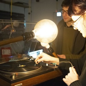 A student places a needle on a record with two large light bulbs in front of her.