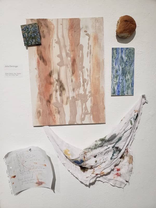 A piece of notebook paper, dinner roll, and pieces of cloth hang on a wall.