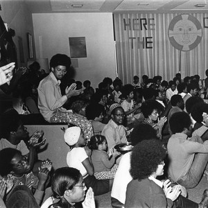 A large group of students wearing afros and 1970s attire sit on the floor and furniture and applaud.