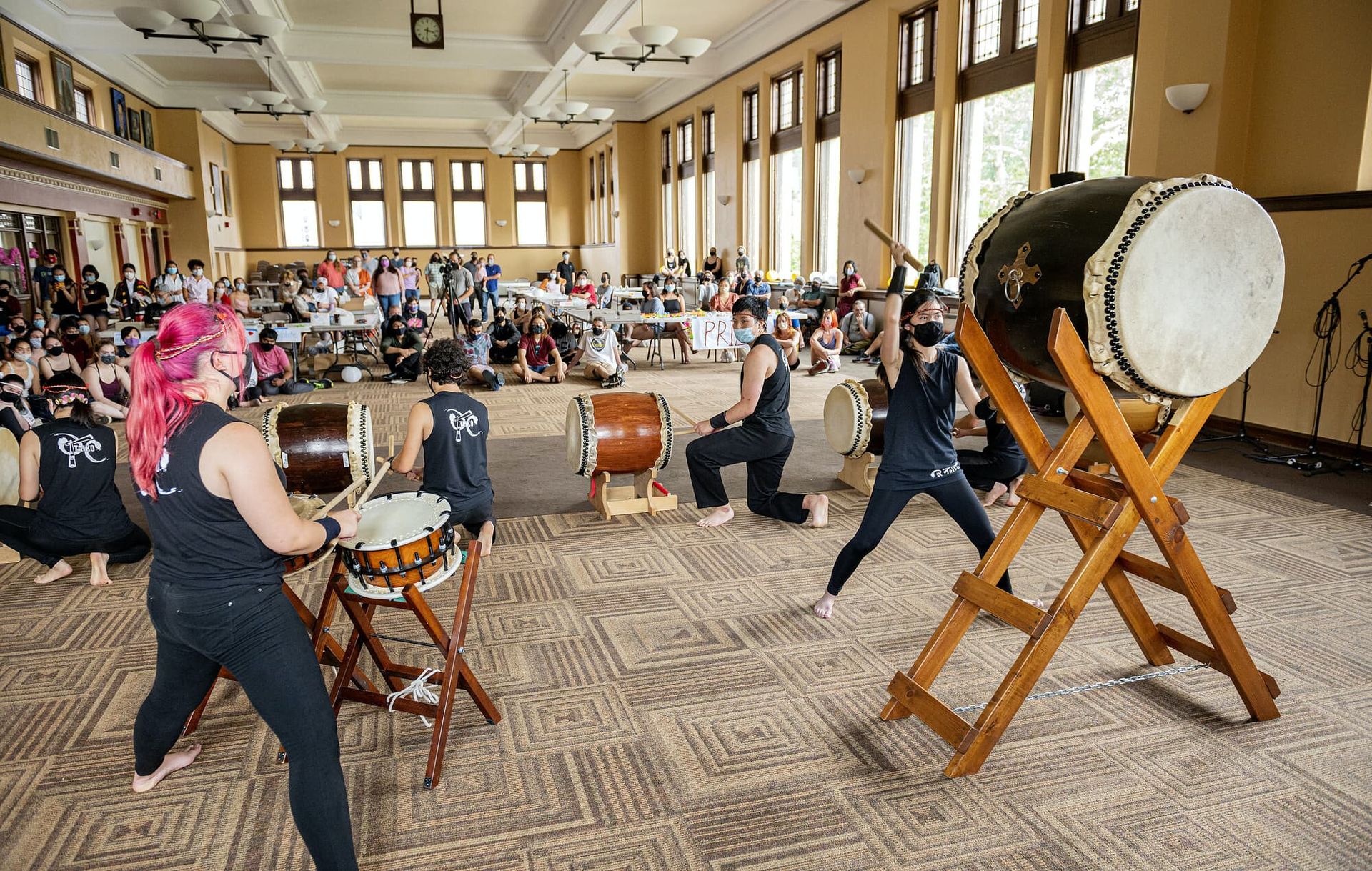 Taiko drummers perform before an audience in a large room.
