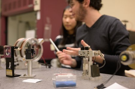 Two students stand in front of a board with physics tools on it. 
