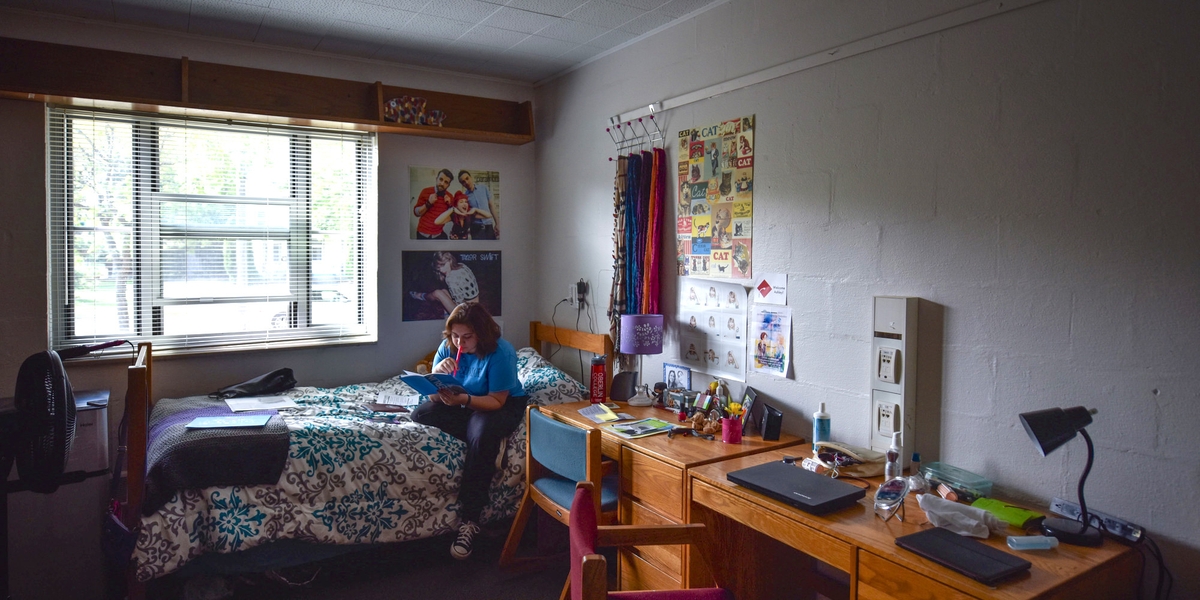 A girl sits on her dorm room bed srrounded by posters on her wall.