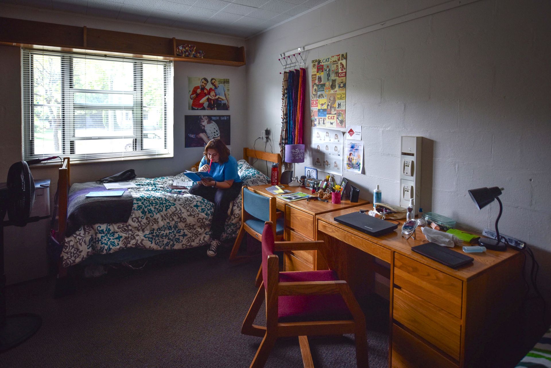 A girl sits on her dorm room bed srrounded by posters on her wall.