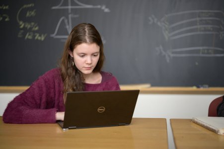 A female student works on a laptop with notes from a physics meeting written on a blackboard behind her.
