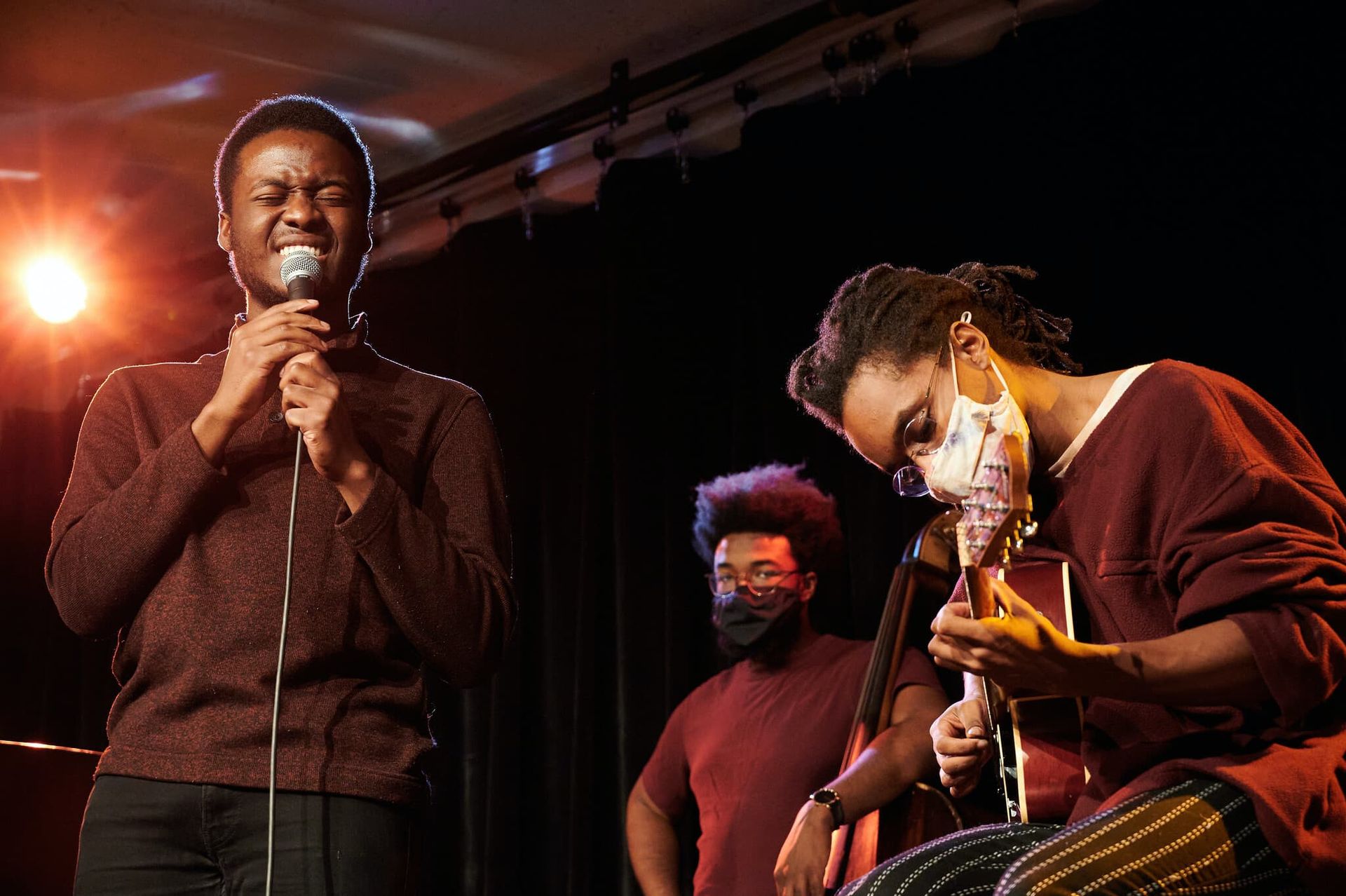 A male student sings into a microphone while two people play instruments next to him.