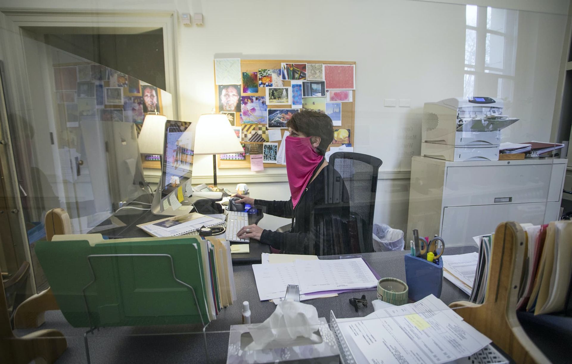 A woman works at her off desk while wearing a face mask and enclosed in plastic barriers.