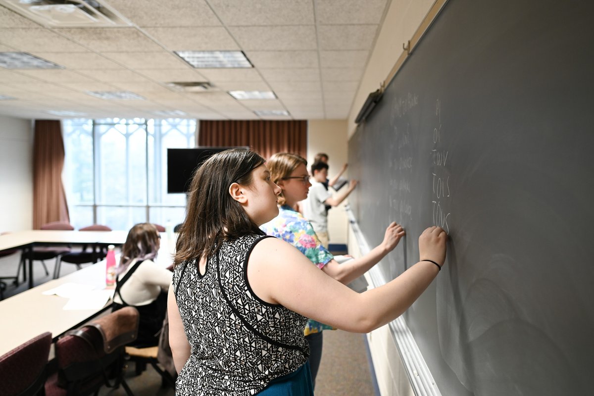 A row of students write Greek on a blackboard.