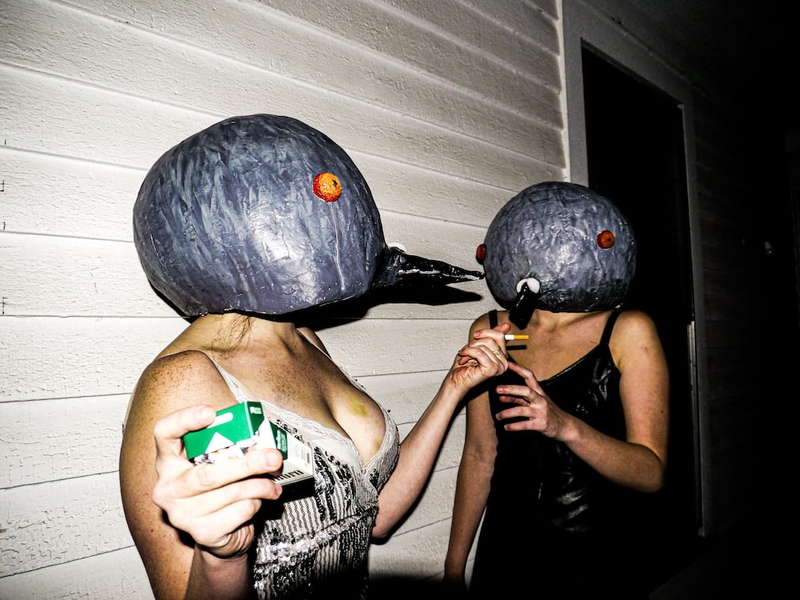 Two people exchanging cigarettes and wearing large paper mache heads with long beaks