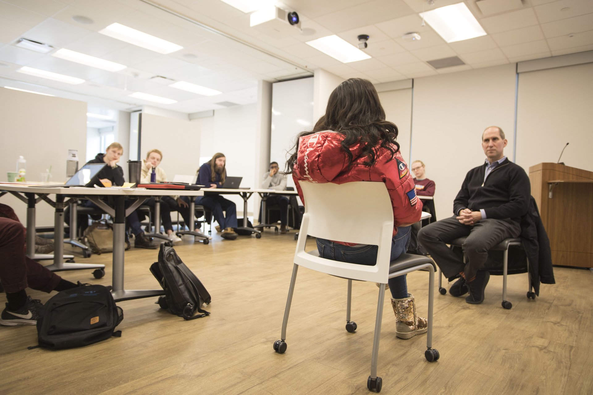 A teacher and student sit across from each other in front of a classroom with students looking on.