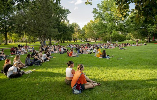 A large group of students sit in a park facing a stage.