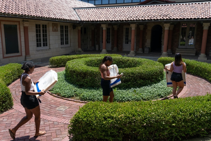 Three students walk through a courtyard carrying large paintings.