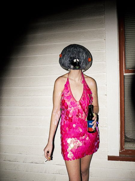A person in a sundress holding a cigarette and bottle, wearing a paper mache head.