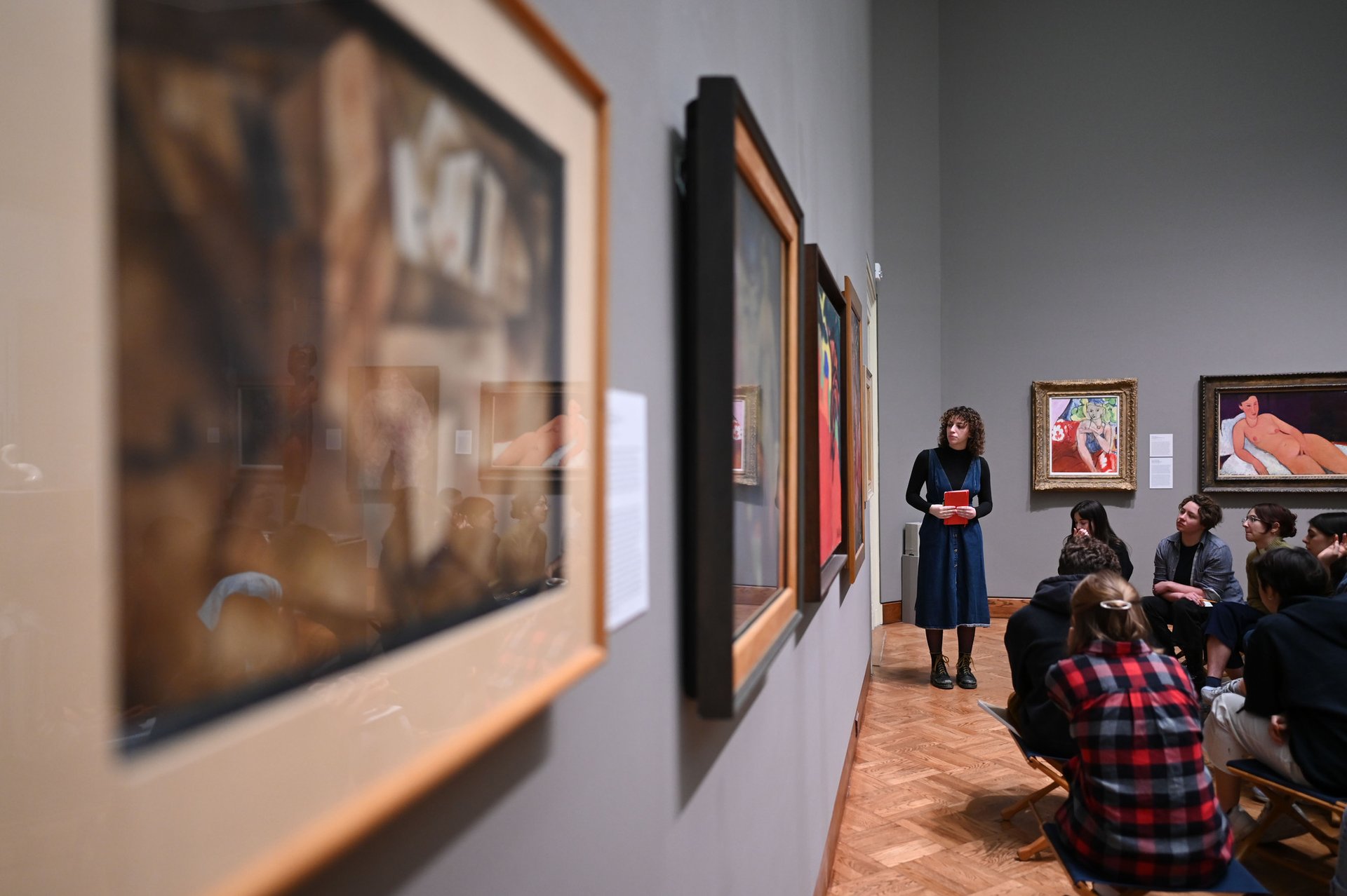 Students sit in an art gallery with another student standing in front of them.